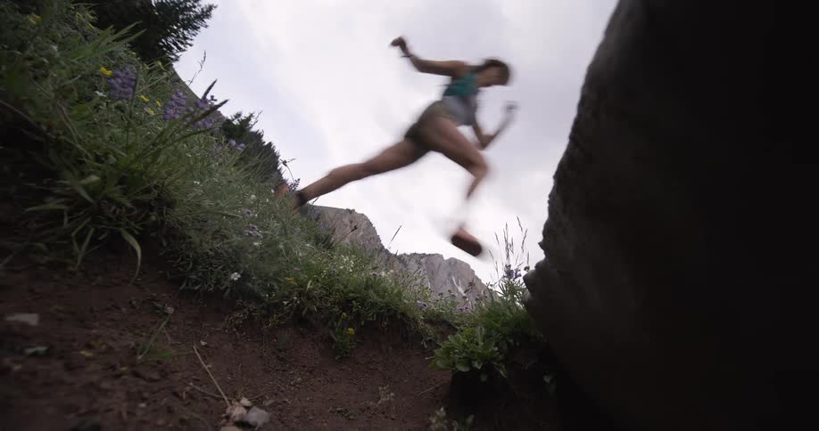 A lowangle view shows the blurry silhouette of a person in athletic clothing leaping between two rocks on a grassy wildflowercovered hill beneath an overcast sky