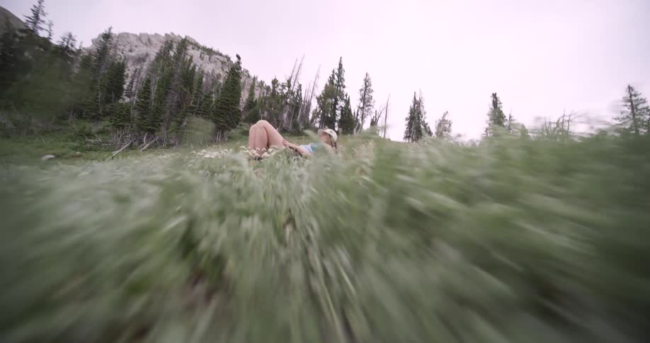 A person wearing a cap and light clothing is first seen sitting then quickly moving to a reclining position among tall green grass and white flowers as the camera rapidly moves forward over the foliage toward them
