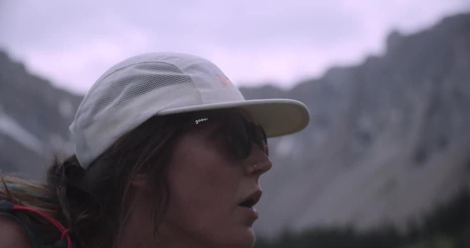 A close view shows a young woman wearing a white cap and sunglasses taking a break from hiking to catch her breath on a green mountainside with rocky peaks visible in the background