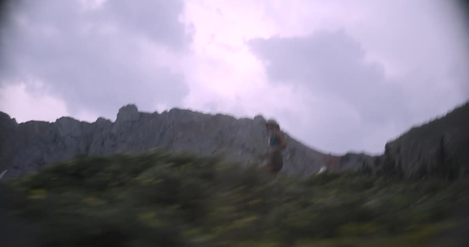 A young woman is along a trail through low green vegetation with a large grey jagged mountain range dominating the background under a cloudy white sky