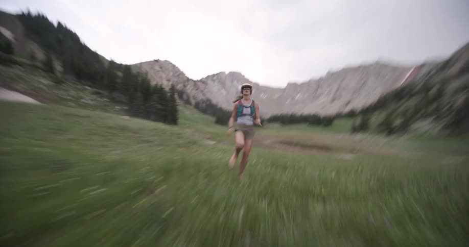 A person runs from the center background toward the camera across a wide green meadow with a small pond and yellow flowers all contained within a deep rocky mountain valley lined with dark pine trees under a white and grey sky