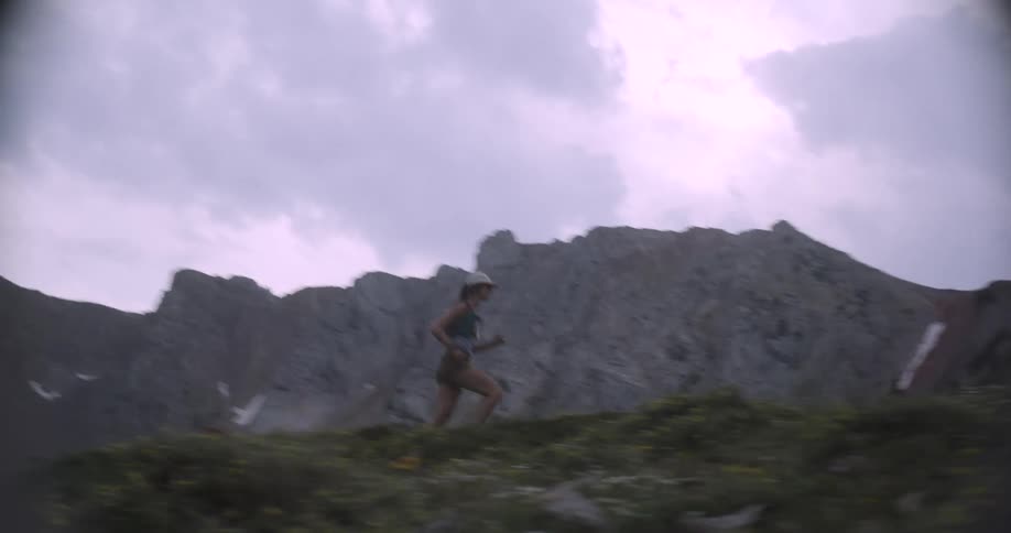 A young woman runs along a mountain trail at midday wearing a hat as the bright sun shines overhead