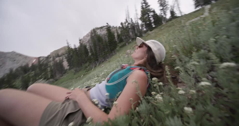 A woman wearing a cap sunglasses and a hydration pack lies down in a grassy field with small white flowers looking up towards the sky with the surrounding mountain scenery out of focus