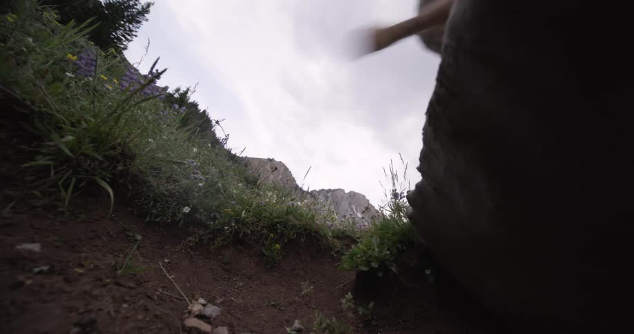 A woman is seen from a low angle running on a rock and grassy path framed by a dark rock mass on the right and low plants with purple flowers on the left set against a background of mountains and an overcast sky