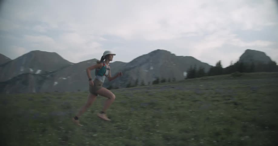 A woman wearing a cap and running gear runs from left to right across a large dark green grassy hillside with a series of grey rocky mountain peaks and valleys in the distant background under a bright overcast sky