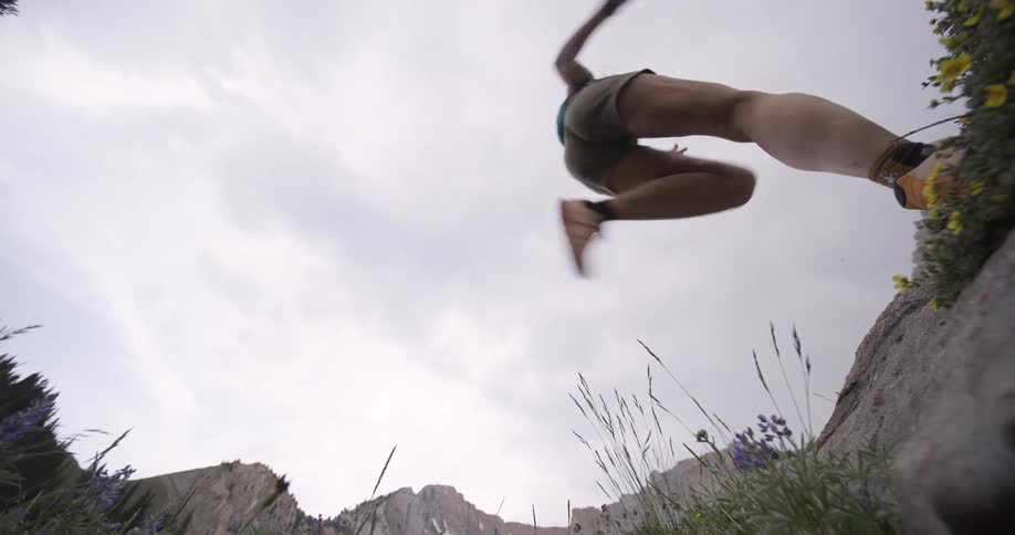 A woman wearing shorts and a vest runs toward the camera from the left with the action viewed from below against a white cloudy sky and a background of dark mountain peaks and small plants