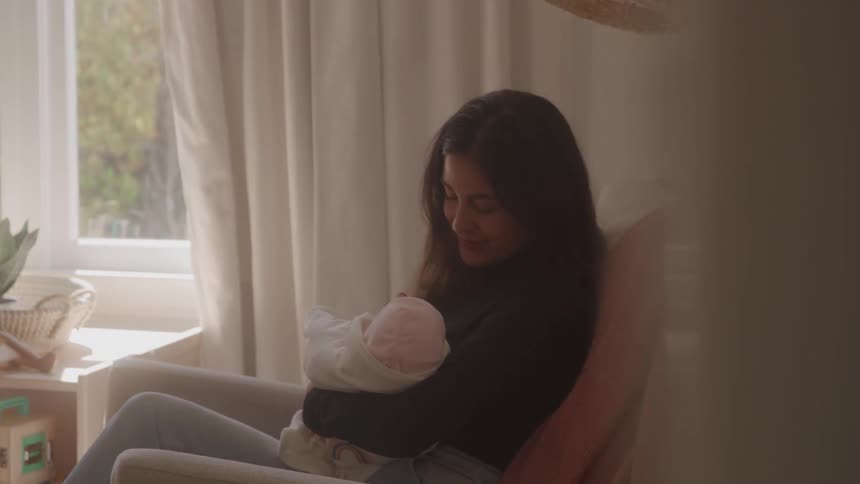 Woman sitting in an armchair holding a baby wrapped in a light blanket beside a window
