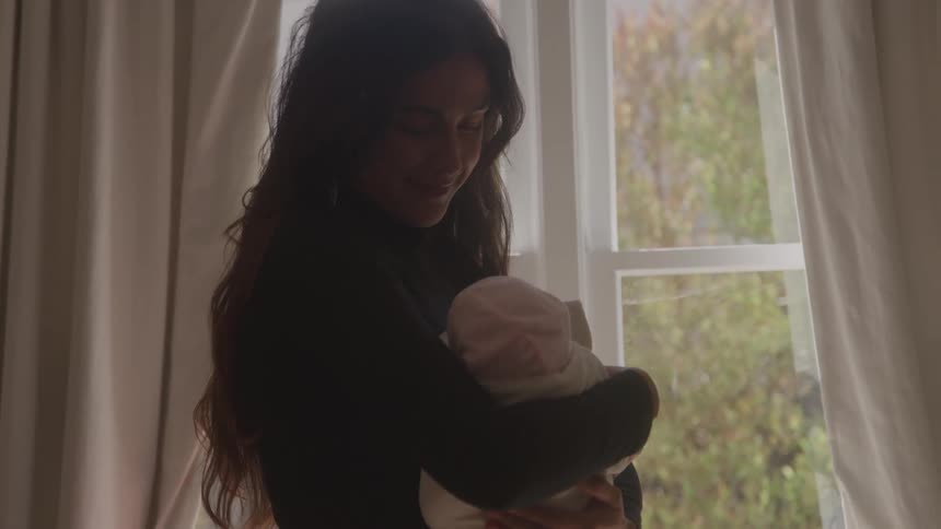 Woman dressed in black holding a baby wrapped in a light blanket near a window