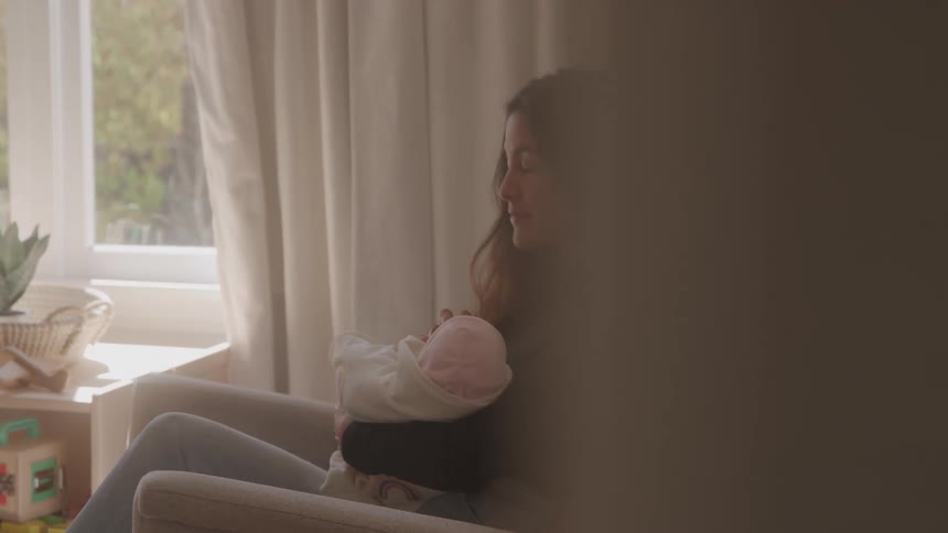 Woman sitting in an armchair holding a baby wrapped in a light blanket while looking at the window