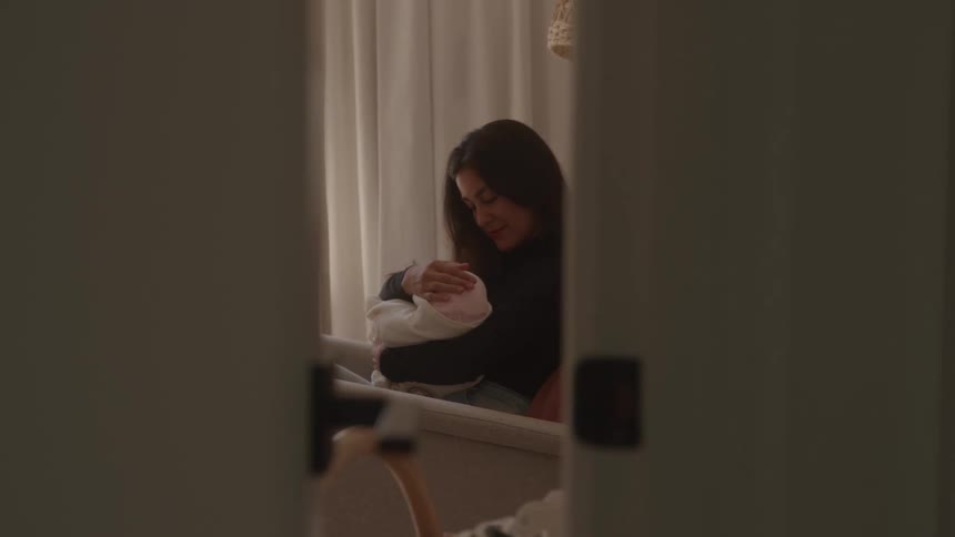 Partial view of a woman sitting and holding a baby wrapped in a light blanket inside a room