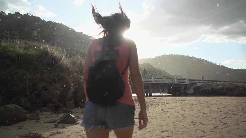 A woman wearing a backpack purse walking towards a bridge and mountains as it sprinkles