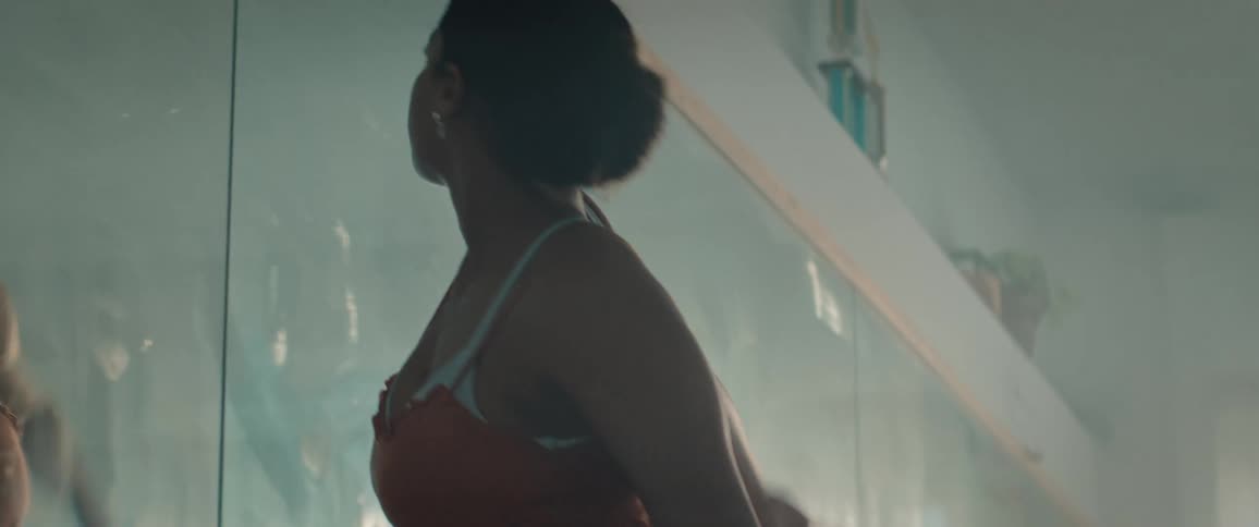 A close view shows the bare feet of a woman on a wooden floor rising onto her toes before she is seen dancing and stretching in a flowing orange dress against a bright windowlit studio