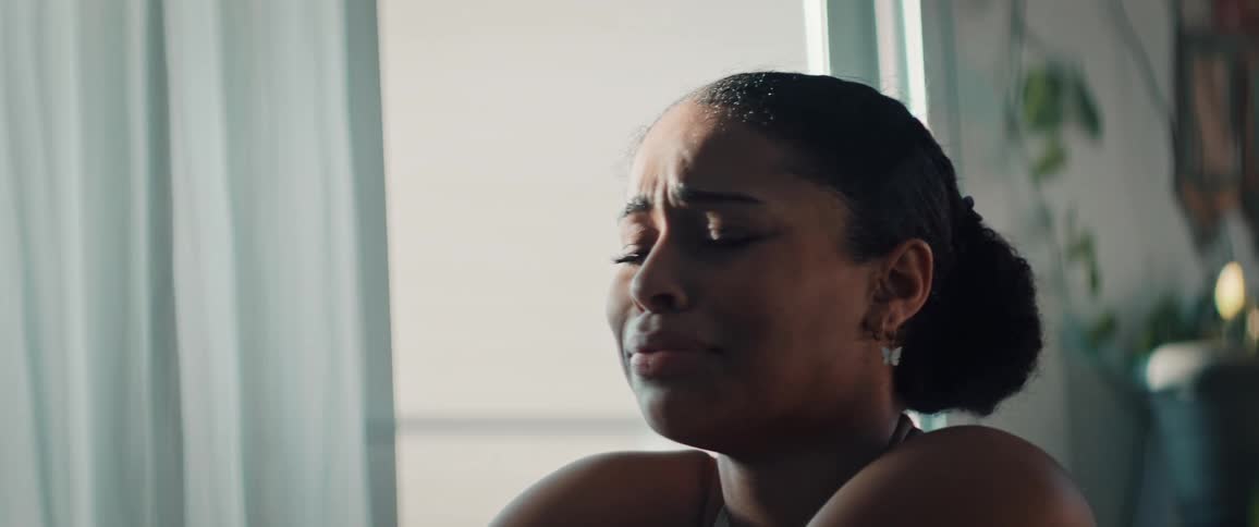 A woman sits by a window crying indoors