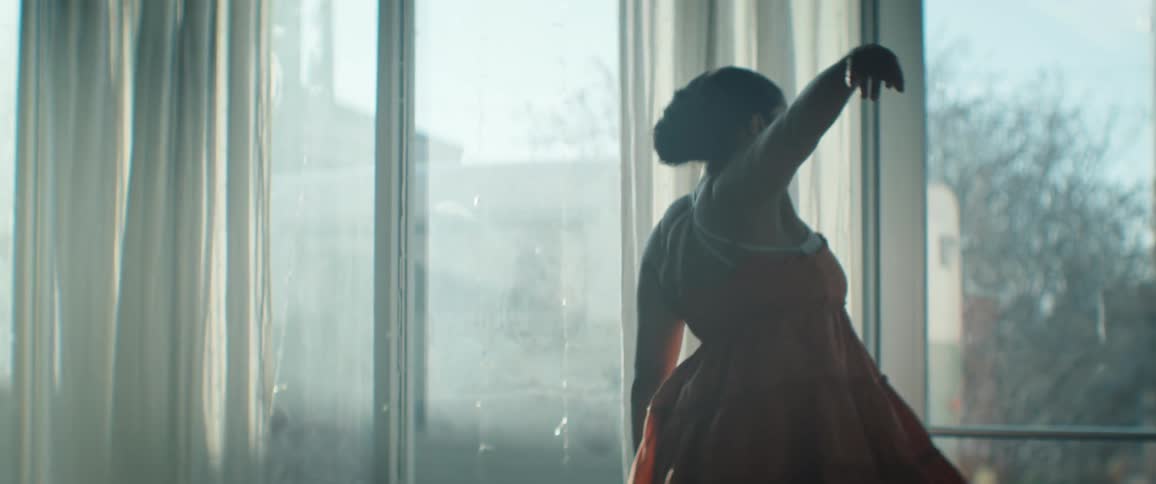 The woman reaches forward and kicks her leg while dancing by the window