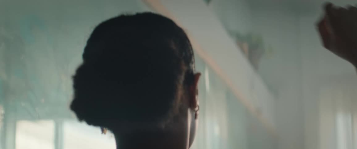 A young woman with dark hair tied up wears a sports bra and stretches her arms looking at her reflection in a bright mirror or glass surface in a moody backlit gym or studio