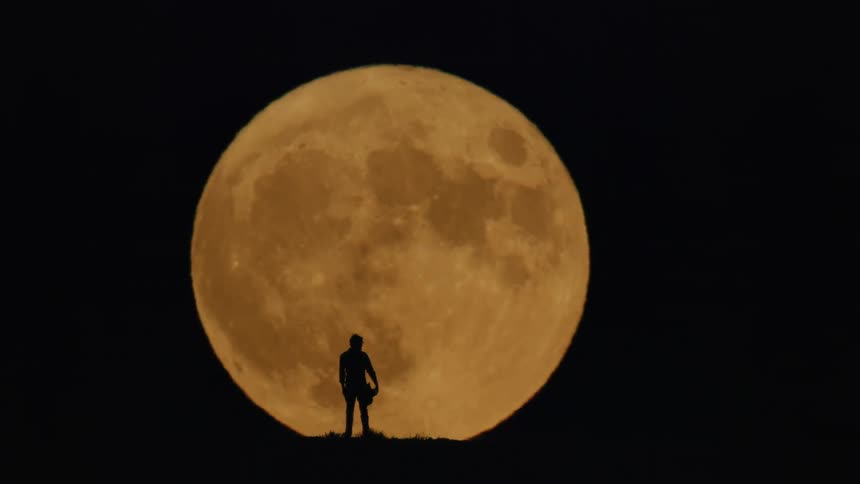 Silhouette of a person wearing a hat in front of the orange full moon in the night sky