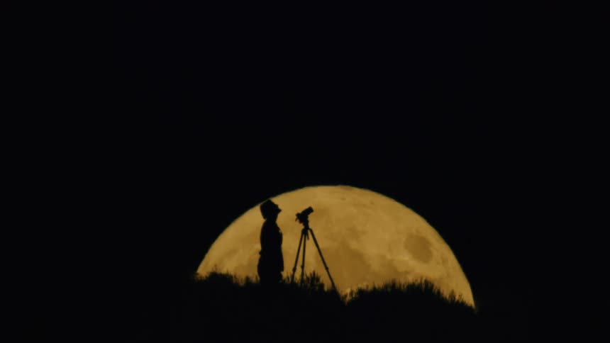 Person standing next to a tripod in front of the moon