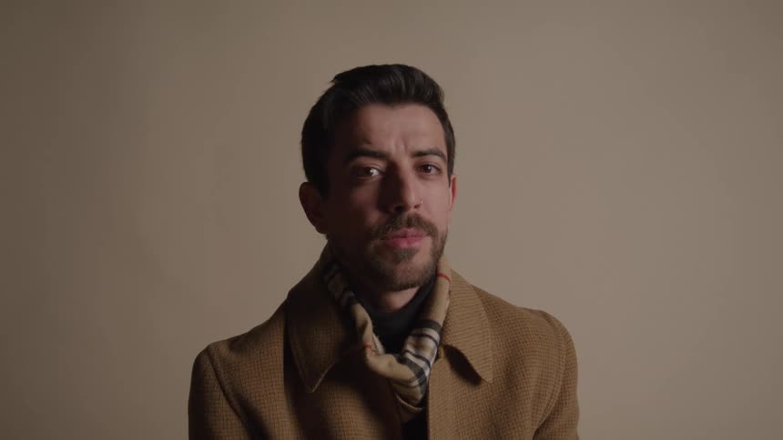 A man with a mustache and beard wearing a brown overcoat and a patterned scarf over a dark turtleneck stands in front of a plain background