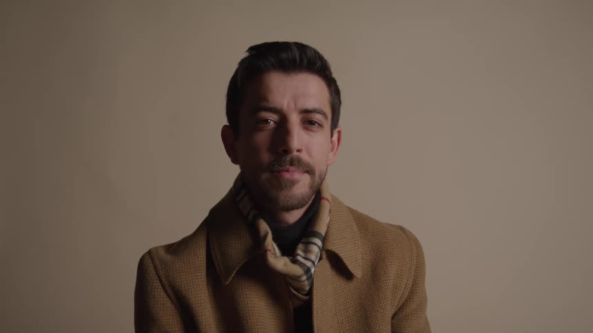 A welldressed man with dark hair and a short beard wearing a tan wool coat and a plaid scarf looks to the side and then directly at the camera with a neutral expression against a plain beige background