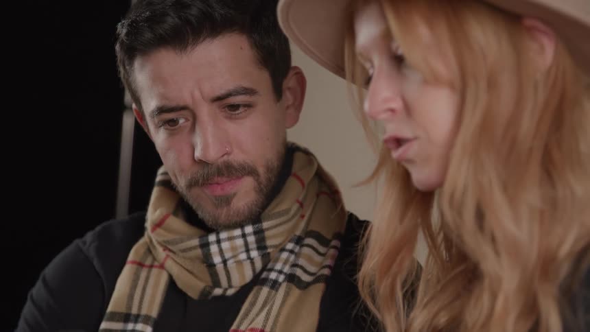 A man in a plaid scarf intently looks at something while a woman with long red hair and a hat speaks to him