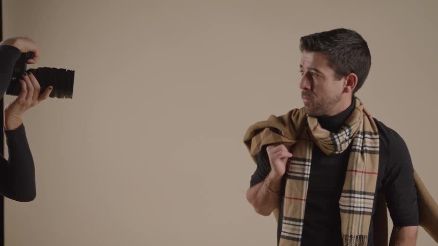 A male model wearing a black t shirt a plaid scarf poses putting a brown coat over his shoulder while a female photographer wearing a fedora shoots picture of him in front of a white background