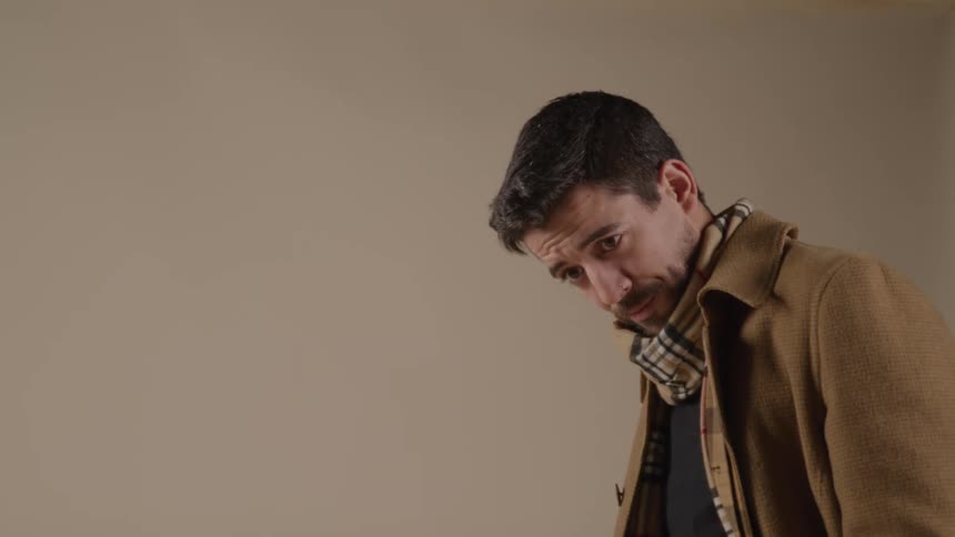 A male model wearing a black shirt a plaid scarf and a brown coat makes poses while a female photographer wearing a fedora shoots picture of him in front of a white background