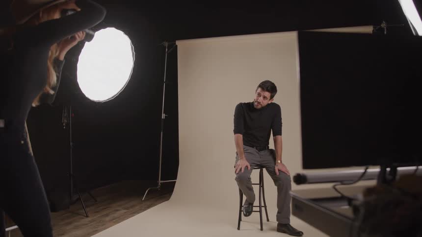 Man sitting on a stool poses while a woman captures shots in the studio
