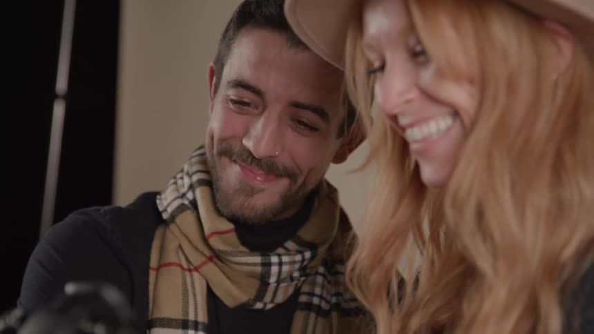 A man wearing a black t shirt and a plaid scarf and a woman wearing a fedora look down at a camera the woman is holding then smile and nod happy with what theyre seeing