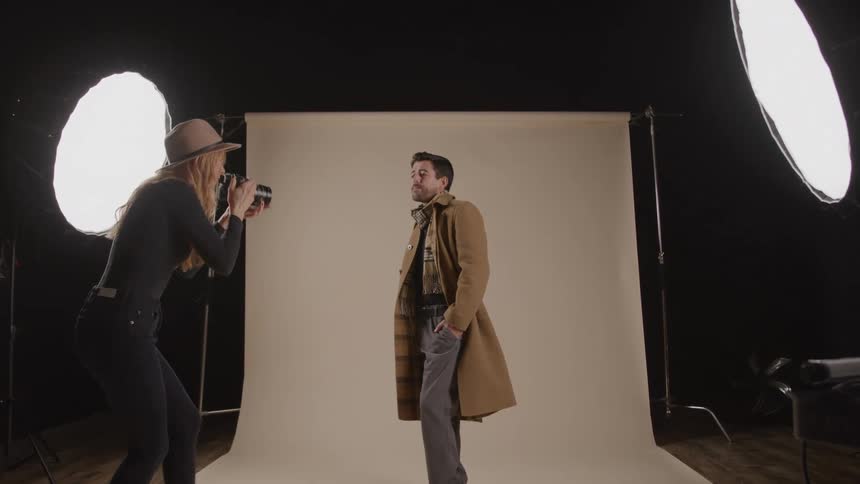 A male model wearing a black shirt a plaid scarf a brown coat and grey pants makes poses while a female photographer wearing a fedora walks around him and shoots picture of him in front of a white background