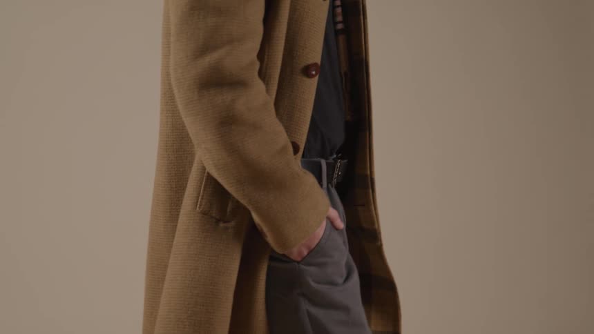 A person in a camel overcoat stands sideon with a hand in their pocket revealing a plaid lining