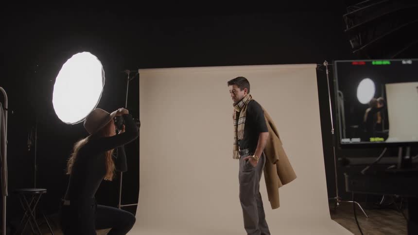 Male model wearing formal clothes posing while a female photographer wearing a hat take pictures of him indoors