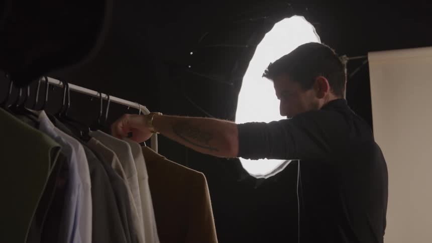 A stylist man wearing a dark turtleneck and gold watch checks a rack of clothing before pulling off a tan coat for a shoot illuminated by a bright professional softbox light in a studio