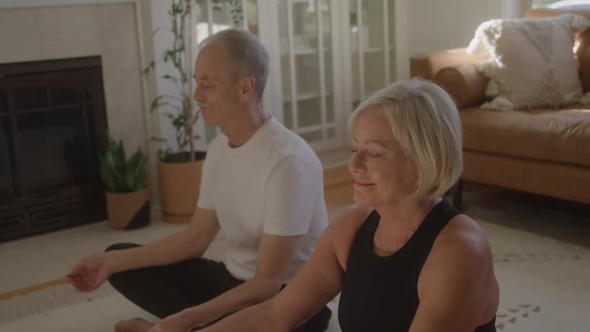 An elderly man sits beside a middleaged woman in deep meditation in the living room at midday