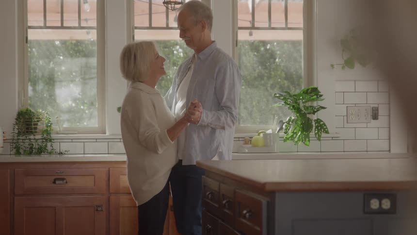 An elderly couple holds hands while dancing in the kitchen at midday their faces glowing with excitement and joy