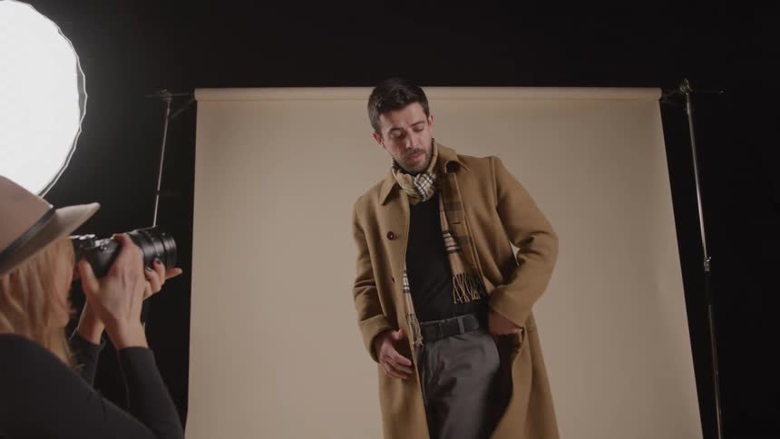 A fashion model man wearing a long tan coat and plaid scarf poses for a photographer woman wearing a hat who captures his photo in a studio setting against a beige backdrop illuminated by bright softbox lights