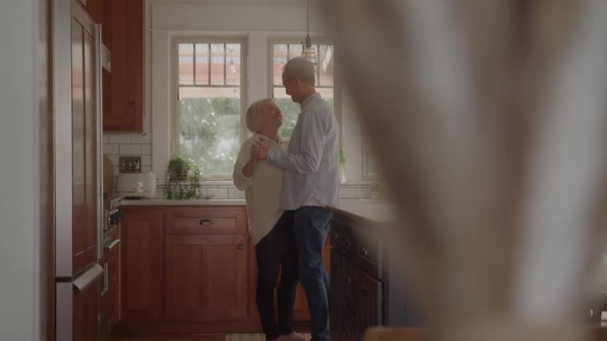 A mature man and woman stand close together in a bright modern kitchen with wooden cabinets and a dark blue island holding hands and doing a slow dancing