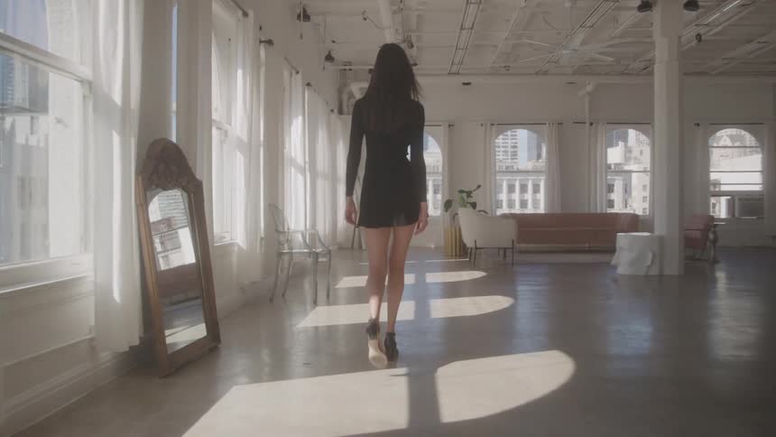 Female model wearing a black dress walking indoors next to a row of windows during day time