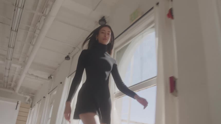 Female model wearing a black dress walking indoors next to a row of windows during day time