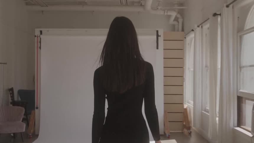 A young woman in a short black dress struts down a studio apartment towards a white screen passing a row of windows with white curtains