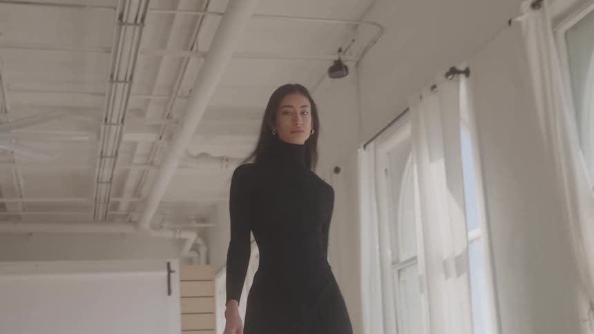 A tall young woman with long dark hair wears a fitted black turtleneck minidress and walks confidently toward the camera in a bright loft studio with white walls large windows and an exposed ceiling