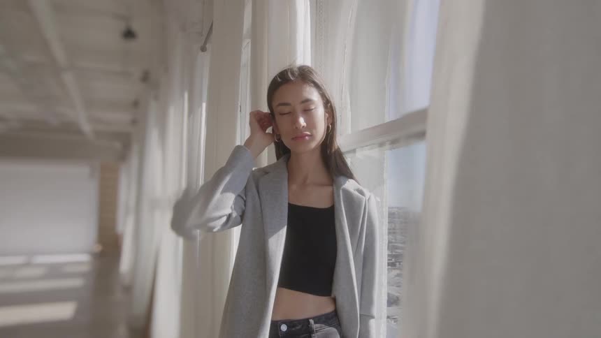 White curtains move with the wind as a young woman wearing a gray coat and a black top tucks her long hair behind her ear