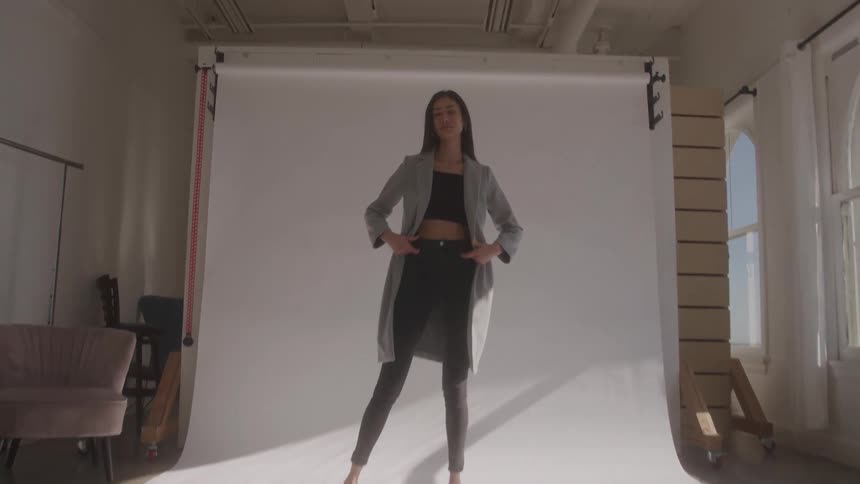 A tall young woman with long dark hair wears a light grey trench coat over a dark crop top and pants while posing confidently on a large white backdrop in a bright photography studio with large arched windows