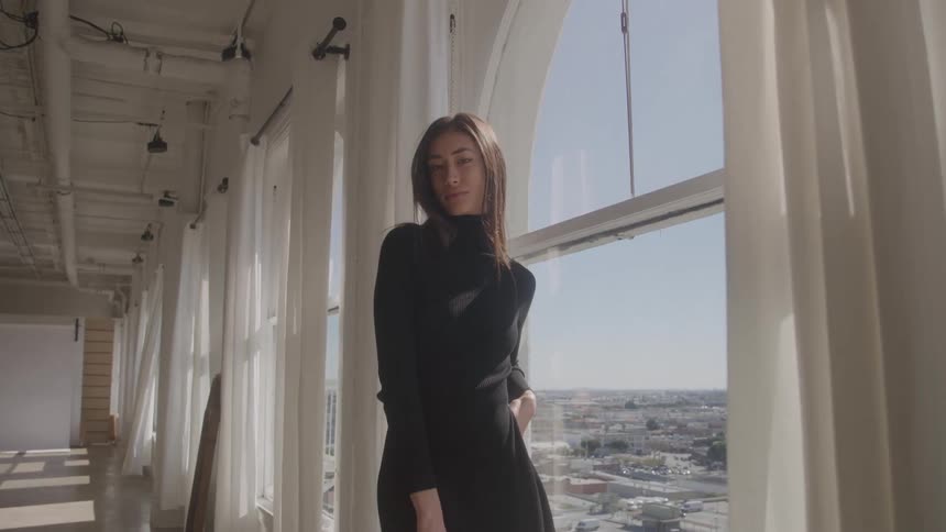 Young adult woman modeling wearing a black dress next to a window indoors during day time