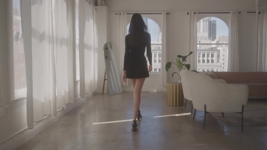 A woman in a black dress and high heels walks away from the camera through a sunlit elegant room with large arched windows overlooking a city