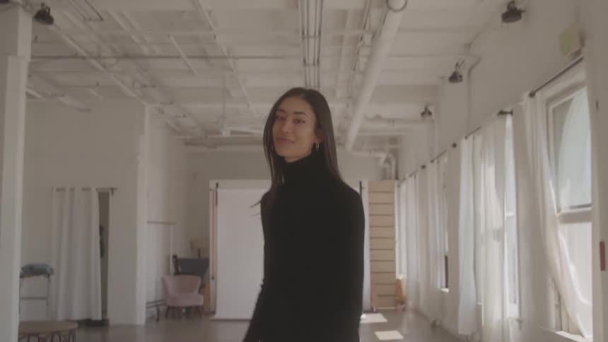 A young woman in a short black dress struts down a studio apartment towards a white screen passing a row of windows with white curtains then turns smiles and without ever stopping turns back in her original direction