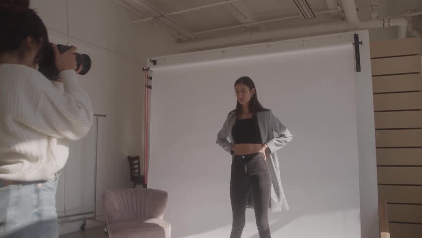 A young woman wearing a gray coat dark gray pants and a black top poses in front of a white background as a photographer takes pictures