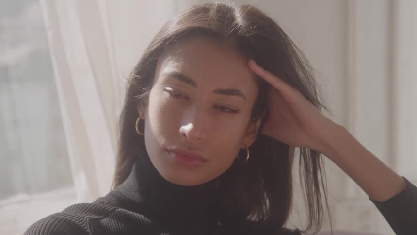 A close portrait view shows a young woman with long dark hair and gold hoop earrings wearing a black turtleneck sweater while gazing seriously at the camera and holding her hand to her head in a bright windowlit room