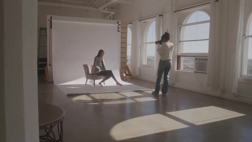 A young woman model wearing dark clothes and a grey jacket sits on a light grey upholstered chair while posing for a photographer man in a bright photo studio with large arched windows and a white backdrop