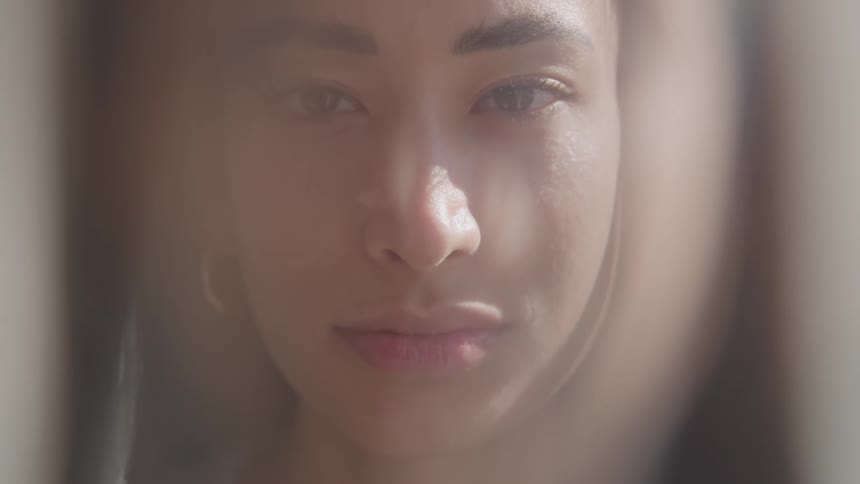 A closeup portrait of a young womans face with dark hair pulled back is captured through a sheer hazy foreground as she opens her eyes to look directly at the camera with a neutral expression in a bright windowlit room
