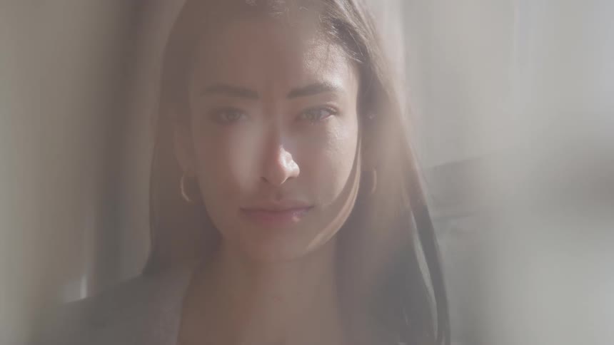 A close portrait view shows a young woman with long dark hair and large hoop earrings looking sadly at the camera through a sheer hazy foreground in a bright softlit room